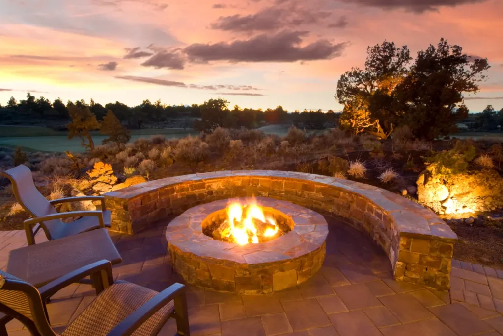 This image captures a stunning outdoor fire pit area set against a picturesque sunset. The fire pit is constructed with natural stone and surrounded by a curved stone bench, providing ample seating. Comfortable chairs are positioned around the fire, creating a perfect spot for relaxation and enjoying the view. The surrounding landscape features native vegetation and adds to the tranquil, natural atmosphere. The warm glow of the fire enhances the beauty of the setting, making it an inviting and serene outdoor retreat.