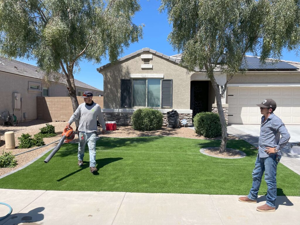 In this photo, two workers are tending to a well-maintained front yard featuring lush artificial grass. The yard is part of a modern home with a beige exterior, neatly landscaped with desert plants and trees. One worker is using a leaf blower to clean the artificial turf, while the other stands with hands on hips, overseeing the work. The house has solar panels on the roof, a stone-accented facade, and a clean driveway. The scene showcases the meticulous upkeep of the artificial grass, highlighting the professionalism and attention to detail of the landscaping team.