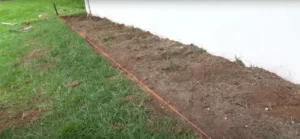 Freshly cleared soil bed alongside a house with a bright orange guideline marking the area for landscaping. Perfect for starting a new garden or laying down edging. Get your outdoor project started today!