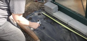 Person carefully cutting holes in landscape fabric with a handheld torch, preparing a precise planting area near a greenhouse bordered by concrete blocks. Perfect for efficient garden setup.